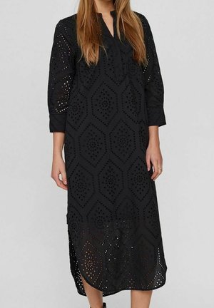 Mujer vistiendo un vestido midi negro de manga larga con patrones geométricos recortados y un cuello, de pie frente a un fondo liso.