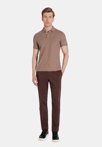 Brązowa koszulka polo z trzema guzikami, w połączeniu z ciemnobrązowymi spodniami. Model nosi czarne trampki z białą podeszwą. Prosty, nowoczesny design.