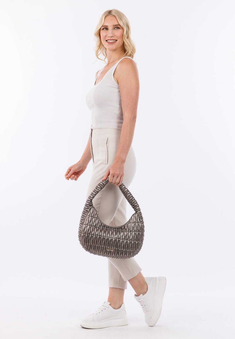 Sac à main texturé gris avec un design tissé, doté d'une poignée épaisse. Une personne portant un haut côtelé et un pantalon beige tient le sac de manière décontractée.