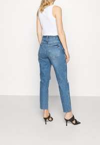 Jeans de mezclilla azul de talle alto con corte relajado, con detalles desgastados en las piernas y dos bolsillos traseros. Combinados con sandalias de tacón negras.