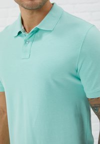 Polo-shirt teal clair en tissu texturé. Présente un col classique et une patte de boutonnage à deux boutons. Manches courtes avec ourlets surpiqués.