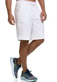 Pantalones cortos de algodón blancos con cinturilla elástica y cordón. Con bolsillos laterales y un ajuste relajado. Usados con zapatillas deportivas grises y azules.