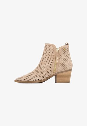 Bota corta de ante beige con perforaciones, punta puntiaguda, cremallera dorada y tacón de madera apilado. Presenta detalles decorativos con tachuelas.