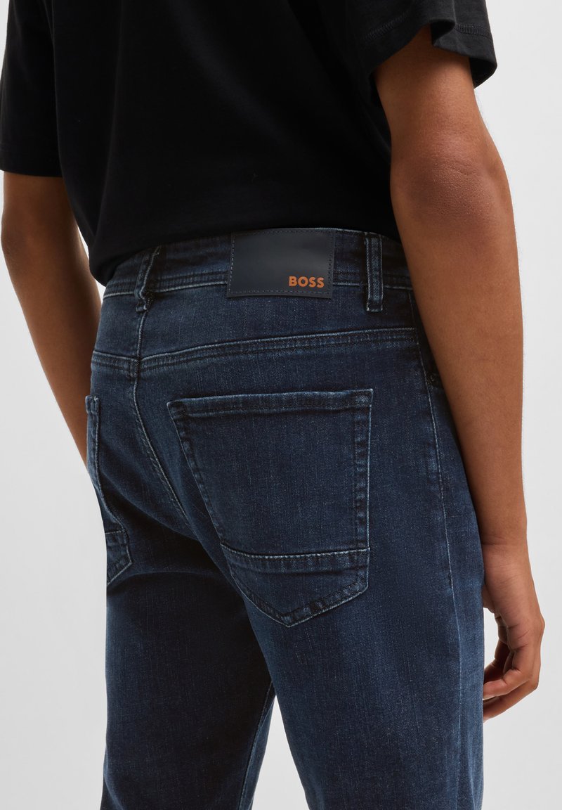 Dunkelblaue Jeans aus Denim haben eine glatte Textur, ein klassisches Fünf-Taschen-Design und einen schwarzen Lederaufnäher auf der Rückseite mit "BOSS" in orangenen Buchstaben.
