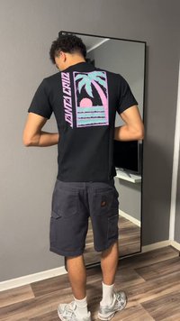 Jeune homme de dos, portant un t-shirt noir avec un motif pastel de palmiers et de coucher de soleil, un short cargo foncé, des chaussettes blanches et des baskets à l'intérieur.