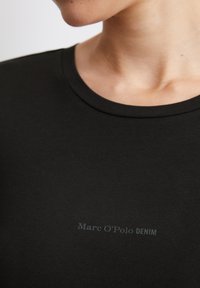 Melns apaļā kakla T-krekls, izgatavots no gludas kokvilnas maisījuma, ar diskrētu logo tekstu "Marc O'Polo DENIM" gaiši pelēkā krāsā priekšpusē.