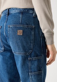Pantalones cargo de mezclilla azul con un bolsillo de parche en la parte trasera, que presenta un parche de logo marrón y detalles de costura visibles.