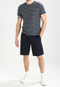Marinblå och vitrandig kortärmad t-shirt som bärs tillsammans med enfärgade marinblå shorts. Modellen har på sig vita sneakers och strumpor som slutar precis vid anklarna, och står framför en enfärgad bakgrund.