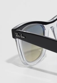 Lähikuva Ray-Ban -aurinkolaseista, joissa on mustat kehysvarret ja kirkkaat kehykset, osoittaen heijastavat gradientti-linssit ja logon tempurissa.