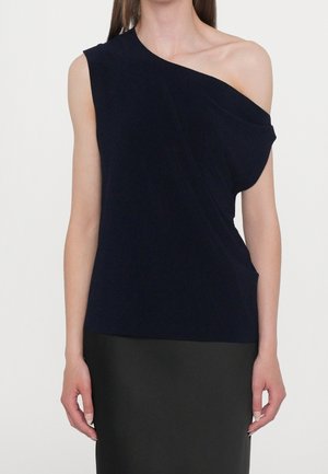 Vrouw draagt een zwarte mouwloze top met één blootgestelde schouder, gecombineerd met een zwarte rok, staand tegen een effen achtergrond.