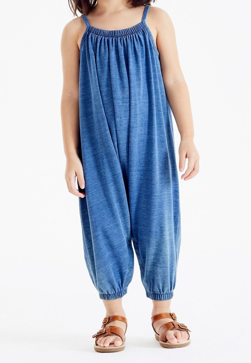 Enfant portant une combinaison bleue ample sans manches avec un col froncé et des sandales marron à boucle, debout sur fond blanc.