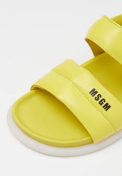 Κίτρινη δερμάτινη σандάλια με δύο φαρδιές λωρίδες, με εκτυπωμένο μαύρο κείμενο "MSGM." Η λεία υφή και η λευκή σόλα τονίζουν τον σχεδιασμό.