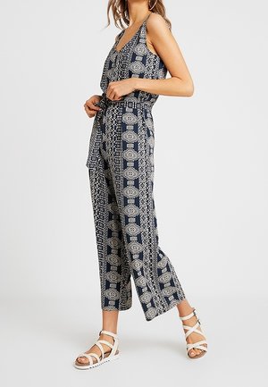 Nainen säätää hihattoman laivastonsinisen haalarin, jossa on valkoisia geometrisia ja kukkakuvioita, yhdistettynä valkoisiin remmihin ja tasapohjaisiin sandaaleihin.