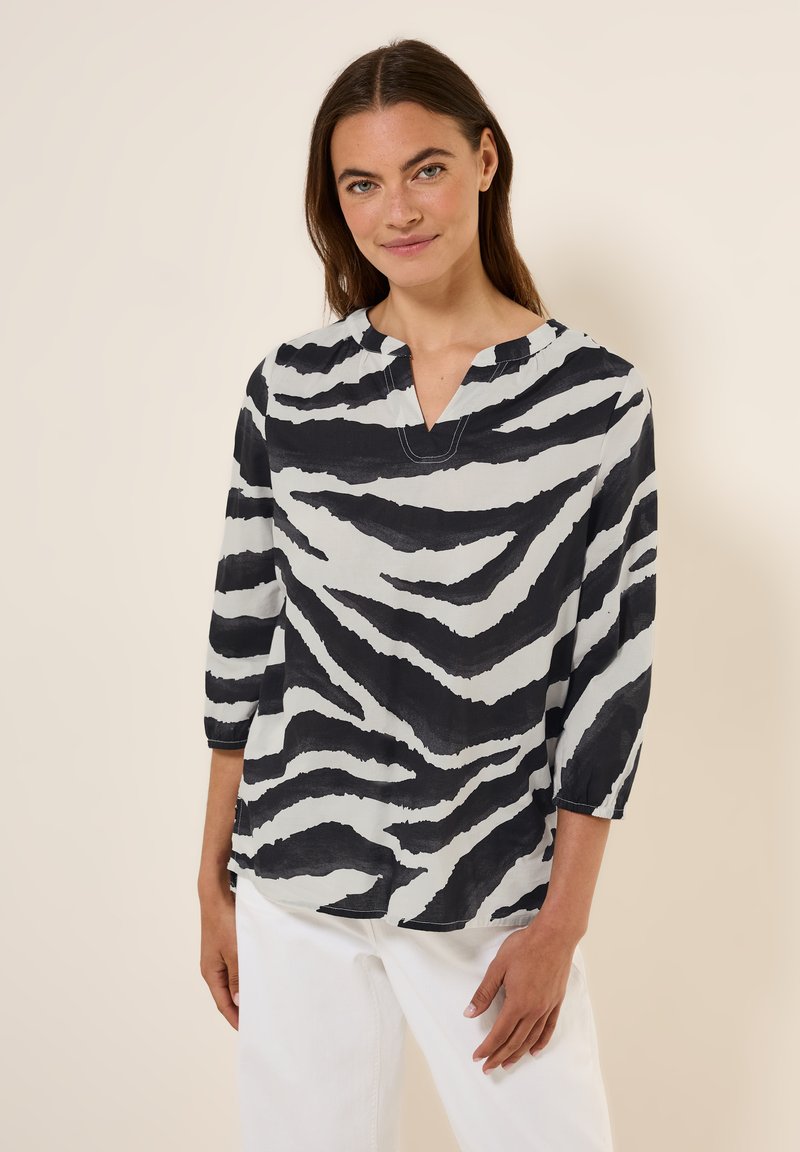 Sort og hvid zebra-stripet bluse med trekvartærmer, V-udskæring og blød, struktureret stof. Kombineret med hvide bukser.
