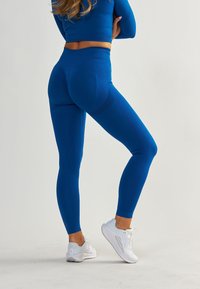 Leggings de sport bleus avec des panneaux texturés, taille haute et design ajusté. Associés à des baskets blanches, vus de l'arrière.