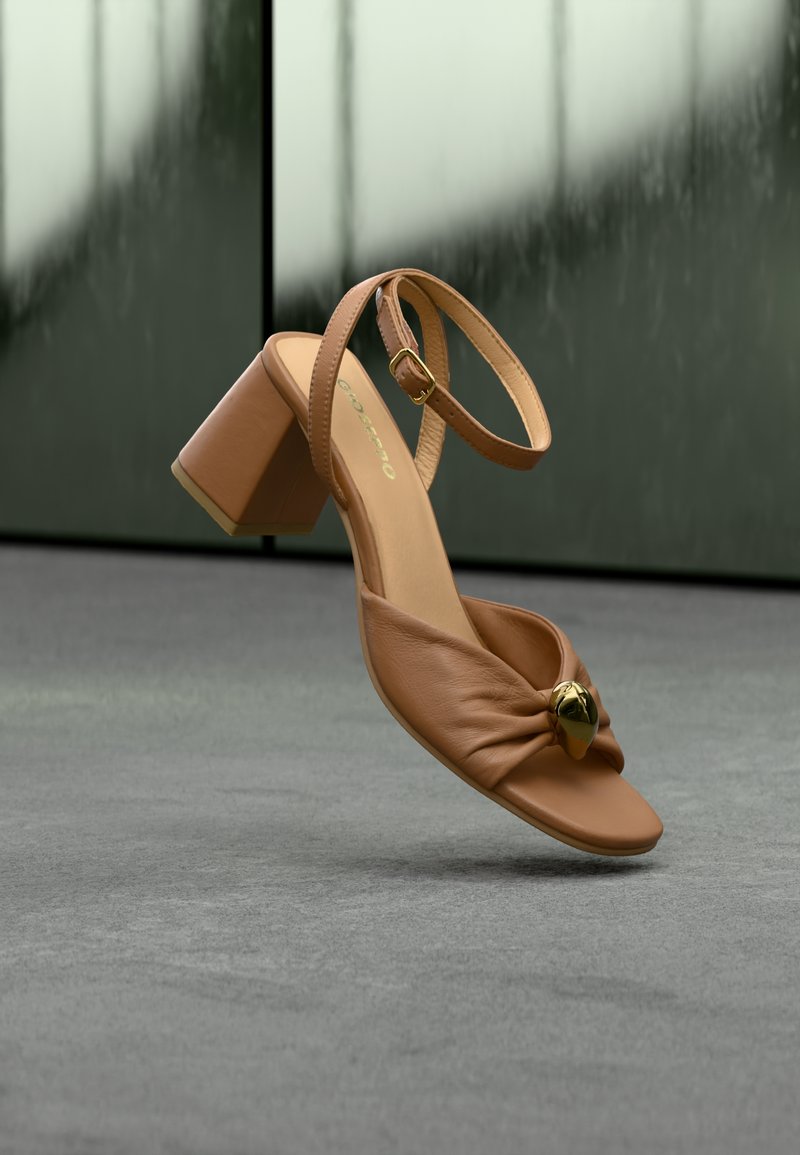 Πέδιλο ανοιχτής μύτης σε ταμπά δέρμα με χοντρό τακούνι, λουράκι στον αστράγαλο, ρυτιδωμένη μπροστινή επιφάνεια και χρυσό διακοσμητικό στοιχείο, τοποθετημένο σε γκρι δάπεδο με θαμπό σκούρο φόντο.