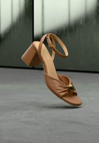 Πέδιλο ανοιχτής μύτης σε ταμπά δέρμα με χοντρό τακούνι, λουράκι στον αστράγαλο, ρυτιδωμένη μπροστινή επιφάνεια και χρυσό διακοσμητικό στοιχείο, τοποθετημένο σε γκρι δάπεδο με θαμπό σκούρο φόντο.