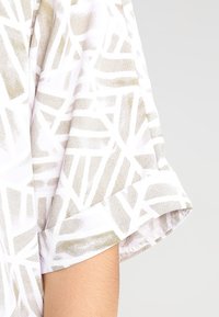 Biała bluzka z geometrycznym złotym i szarym wzorem, z podwiniętymi rękawami. Lekki materiał o delikatnej strukturze i luźnym kroju.