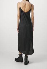 Robe maxi noire sans manches avec un dos en V profond, fabriquée en tissu lisse et léger ; portée avec des bottes de combat noires épaisses.