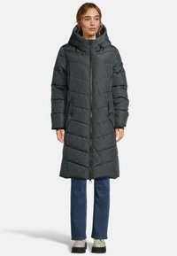 Femme portant un long manteau d'hiver noir rembourré avec capuche, un jean bleu et des chaussures gris clair, se tenant devant un fond blanc uni.