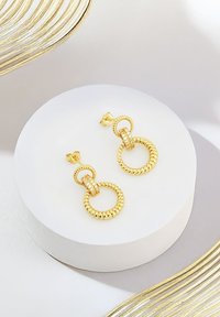 Goldene Creolenohrringe mit strukturierten und glatten runden Gliedern, die mit kleinen klaren Edelsteinen am Verbindungsglied akzentuiert sind. Auf einem weißen Podest ausgestellt.