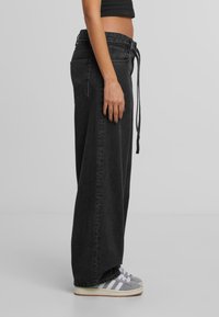 Henkilö, jolla on yllään löysät mustat farkut, joissa on nyöri vyötäröllä, lyhyt musta toppi ja harmaat Adidas-tennarit valkoisilla raidoilla.