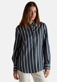 Noor naine, kes kannab tumesinist särki valgete vertikaalsete triipudega ja kreemivärvi pükse, seisab ühetaolise heleda tausta vastu.