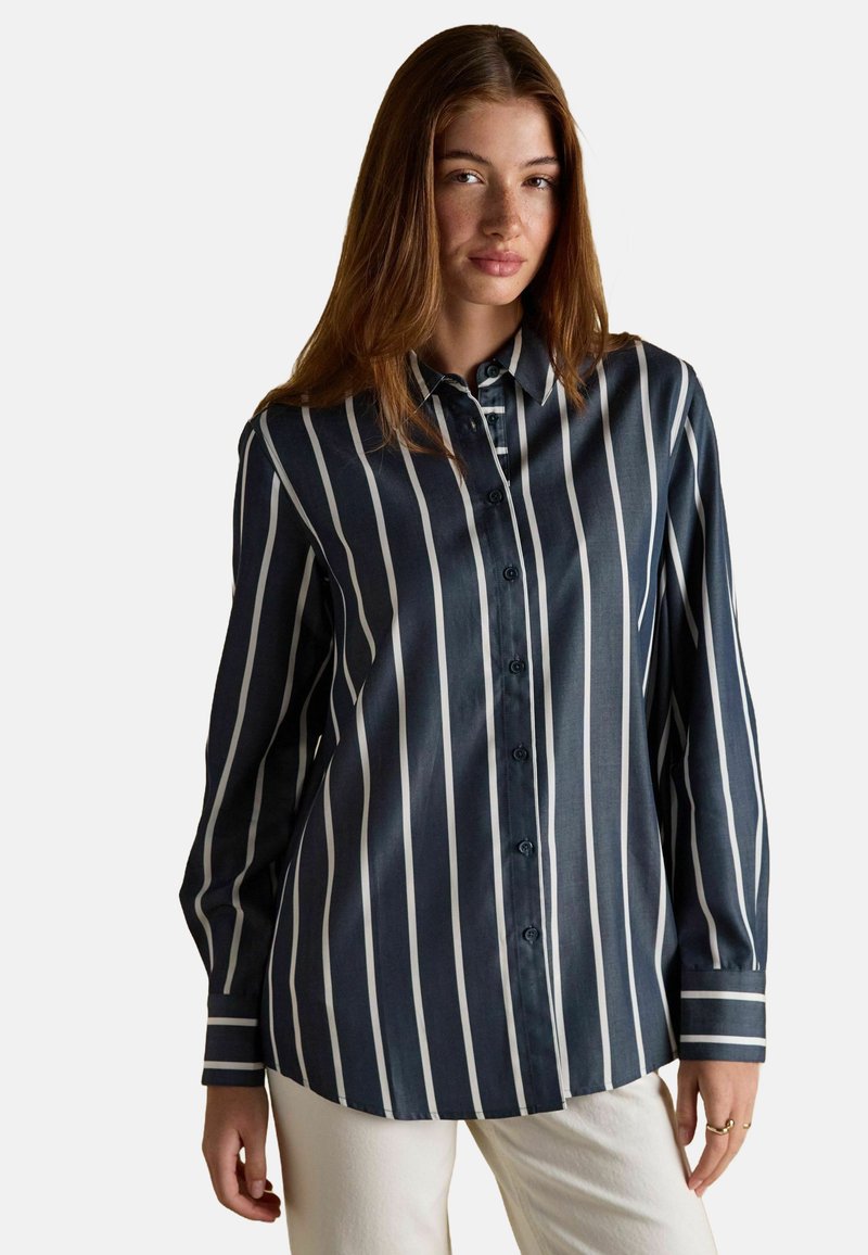 Noor naine, kes kannab tumesinist särki valgete vertikaalsete triipudega ja kreemivärvi pükse, seisab ühetaolise heleda tausta vastu.