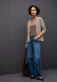 Beige cardigan s otevřeným předním dílem přes hnědý top, doplněný světlomodrými širokými džíny a černými špičatými botami, doplněný hnědou kabelkou.