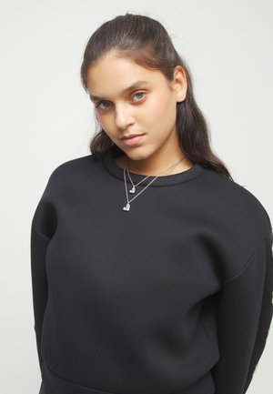 Jeune femme aux cheveux foncés attachés, portant un sweat-shirt noir et deux colliers argentés en forme de cœur, regardant légèrement vers le bas.