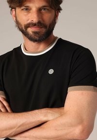 T-shirt noir à manches courtes, avec une bordure en mesh beige et un logo métallique sur la poitrine. Texture lisse et encolure ronde.