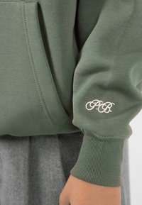 Manga de sudadera verde con las iniciales bordadas en blanco "PB" sobre el puño acanalado, y un bolsillo frontal visible cerca de la muñeca.