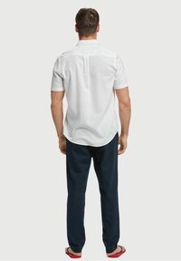 Bílá lněná košile s krátkými rukávy a zadním sedlem, doplněná tmavě modrými kalhotami a červenými žabkami. Hladká textura a pohodlný střih.