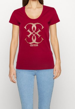 Vrouw draagt een nauwsluitend rood T-shirt met korte mouwen en een gouden, met patroon versierde "GUESS"-logo op de borst, gecombineerd met lichtblauwe jeans.