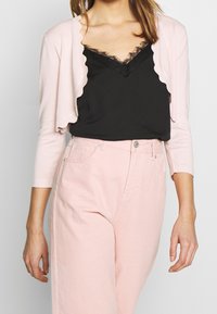 Ljusrosa cardigan med vågiga kanter, som bärs över en svart cami med spetsdetaljer, matchad med pastellrosa högmidjade byxor.