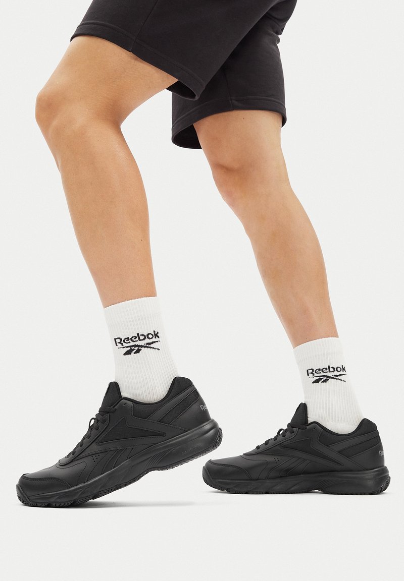 Pantofi sport negri cu un superior sintetic neted, guler căptușit și talpă texturată. Purtați cu șosete albe cu logo-ul Reebok.