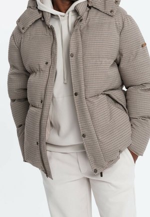 Osoba nosząca beżowo-brązową pikowaną kurtkę w kratę na jasnoszarej bluzie z kapturem i dopasowanych jasnoszarych spodniach, z rękami w kieszeniach.