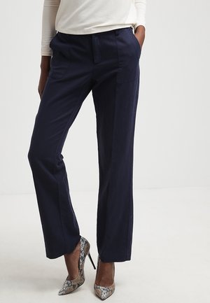 Person trägt marineblaue, gerade geschnittene Hose, weißes Langarmshirt und High Heels mit Schlangenmuster, steht mit einer Hand in der Tasche.