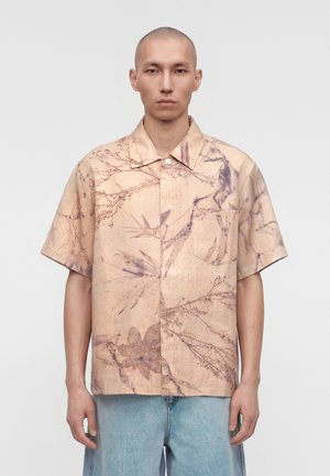 Άνδρας που φοράει μπεζ κοντομάνικο πουκάμισο με βοτανικό σχέδιο και ανοιχτό μπλε τζιν, στέκεται μπροστά σε απλό λευκό φόντο.