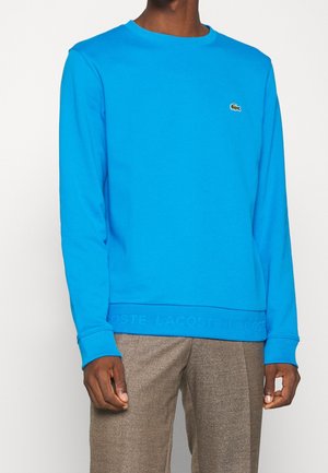 Homme portant un sweat-shirt Lacoste bleu vif à manches longues avec un pantalon marron, debout devant un fond clair uni.