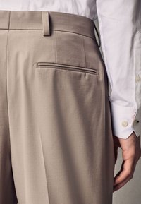 Beżowe spodnie materiałowe mają subtelną fakturę i pojedynczą tylną kieszeń z kontrastowym szwem. Biała koszula jest częściowo widoczna.