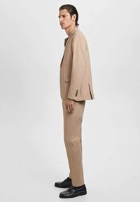 Man in tan suit and black dress shoes standing in profile against a plain light background.