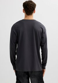 Hombre con cabello corto y rizado, vistiendo una camiseta de manga larga oscura y lisa y pantalones cargo negros, de pie frente a un fondo claro y sencillo.