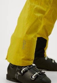 Pantaloni de schi impermeabili galbeni, cu o textură netedă, având un tiv cu fermoar și un detaliu cu logo pe lateral. Sub ei sunt purtate cizme de schi negre.
