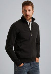 Chaqueta negra con cremallera y hombros y cuello texturizados, cuenta con bolsillos laterales, puños acanalados y un diseño ajustado.