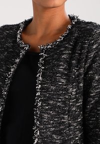 Veste en tweed noir et blanc avec tissu texturé, bords effilochés et design à ouverture frontale. Présente un col rond et un léger éclat.