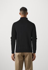 Schwarzer Strickpullover mit hohem Kragen, gerippter Textur und schmalen Ärmeln. Getragen mit beigen Hosen, zeigt ein einfaches, modernes Design.