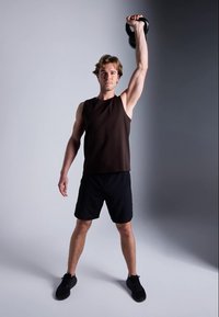 Homem com uma t-shirt preta e calções levanta um kettlebell acima da cabeça com o braço direito, de pé sobre um fundo cinza em duas tonalidades.