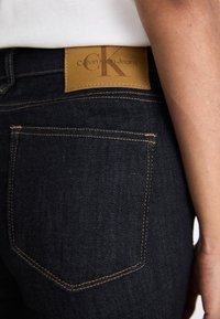 Temno jeans jakne z usnjenim označevalcem s povdarenim napisom "Calvin Klein Jeans", rumeno-oranžnimi šivi ob žepih in prilagojenim krojem.