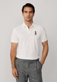 Biela polo košeľa s kontrastným žltým golierom, na hrudi s malým vyšívaným motívom a logom, priliehavý strih, krátke rukávy.
